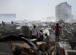 A scene of devastation following an earthquake - piles of rubble and collapsed buildings. Several people are visible searching through the rubble