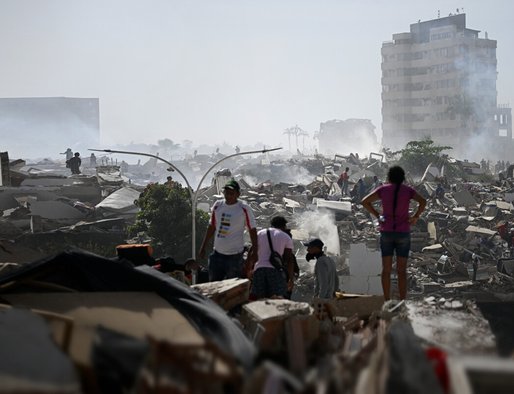 A scene of devastation following an earthquake - piles of rubble and collapsed buildings. Several people are visible searching through the rubble