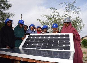 A group of 8 women wearing hard hats stand around a solar panel