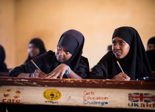 Two girls sitting behind a school desk taking notes. Logos for CARE, MDFHS, Girls' Education Challenge and UK Aid are on the desk