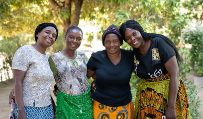 A group of four smiling women pose for the camera together underneath a tree