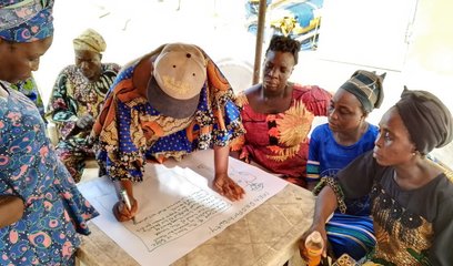 A group of six women gather round a table, one of the women is writing on flipchart paper
