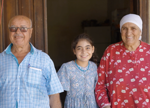 An older womand man stand smiling in a doorway with a young girl between them, who looks like she may be their grand-daughter or another young relative