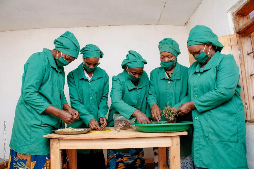 A group of five women in green smocks gather round a table sorting through spices together