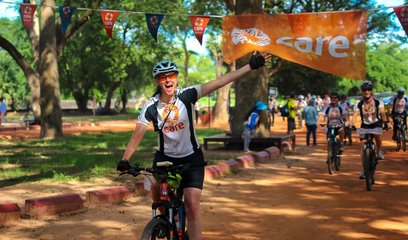 A woman crosses a finish line on a bike with her left arm raised in celebration. She is wearing a T shirt with a CARE logo and there are other cyclists in the background