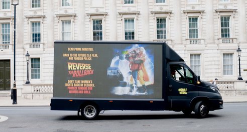 A truck drives in front of a UK parliamentary building with the slogan 'Dear Prime Minister, Back to the Future is a film, not foeign policy Reverse the Rollback. don't take women's rights back by decades. Protect funding for women and girls.